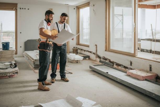 Two men stand in an unfinished house, reviewing construction plans and discussing the details of a home remodeling project.